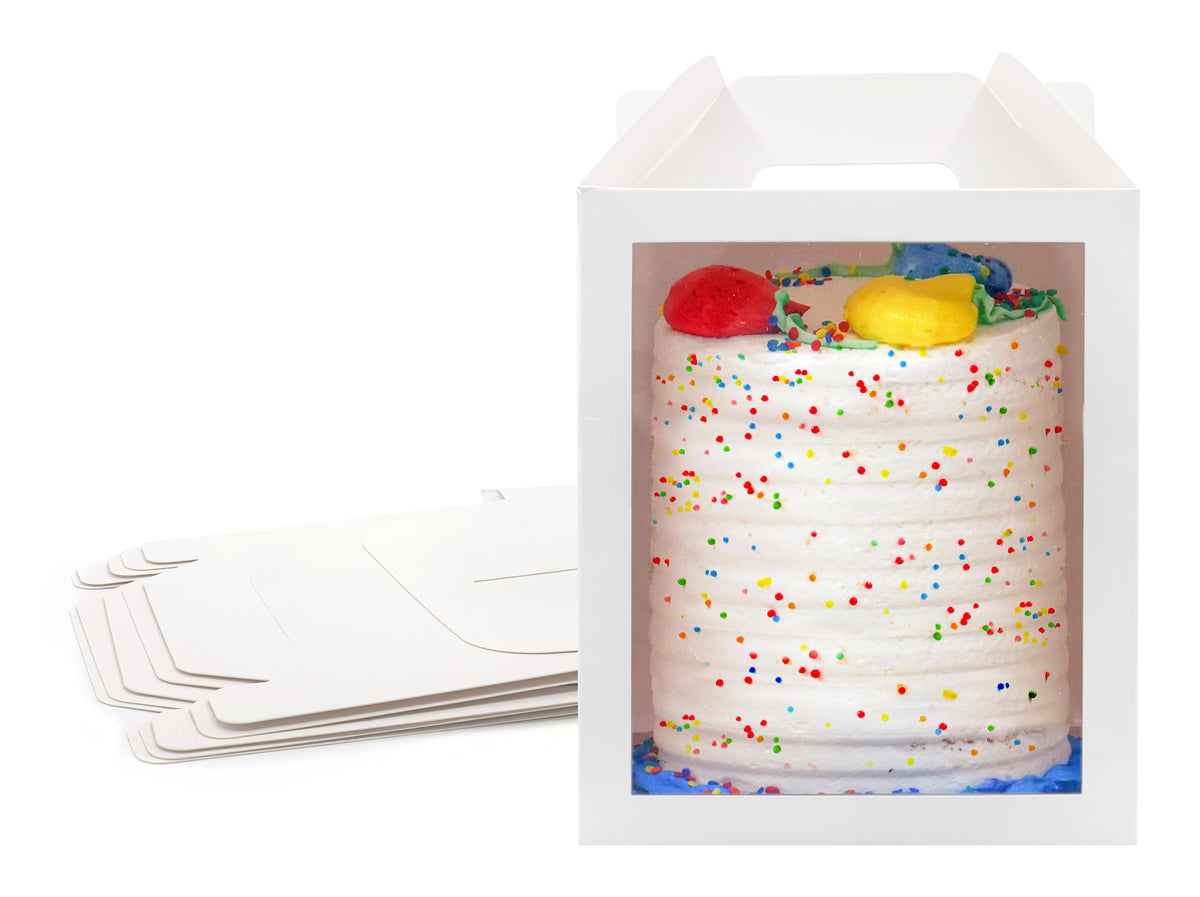 A cake in a 10x10x12 white paper cake box with window and handle, with a stack of folded boxes in the background