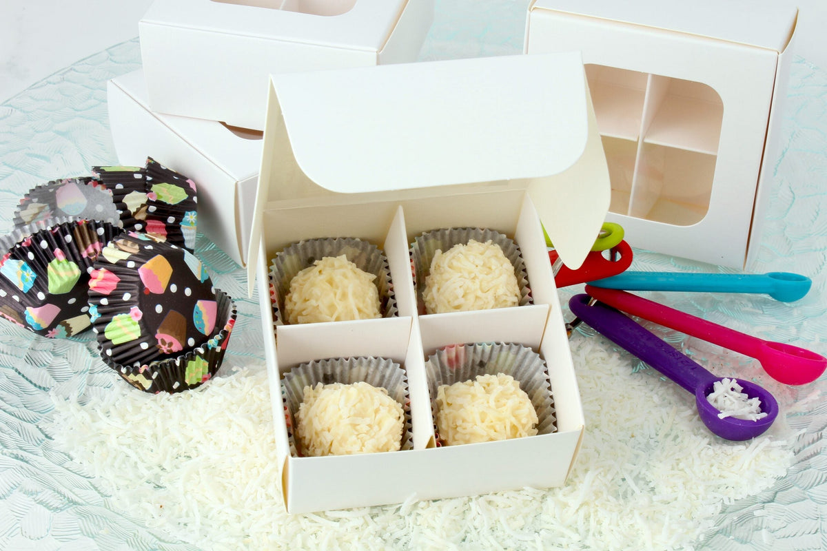 Open white treat box with a four-compartment divider holding coconut truffles, folded boxes behind