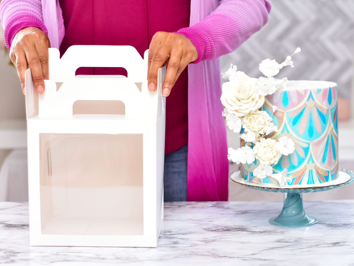 Hands holding the top of a folded 8 by 8 by 8 inch white cake box, a decorated cake beside it
