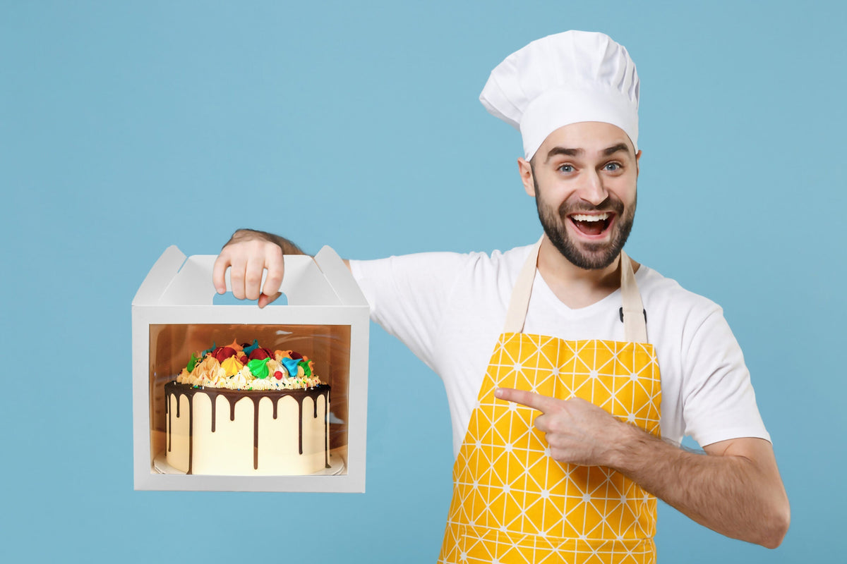 A baker holding a decorated cake in a white cake box with window and handle