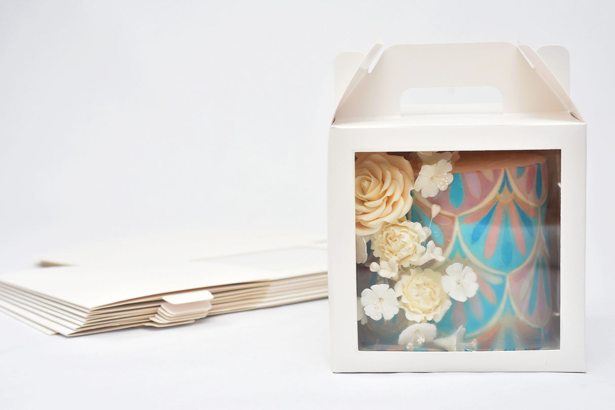 A cake in a 8x8x8 white paper cake box with window and handle, with a stack of folded boxes in the background