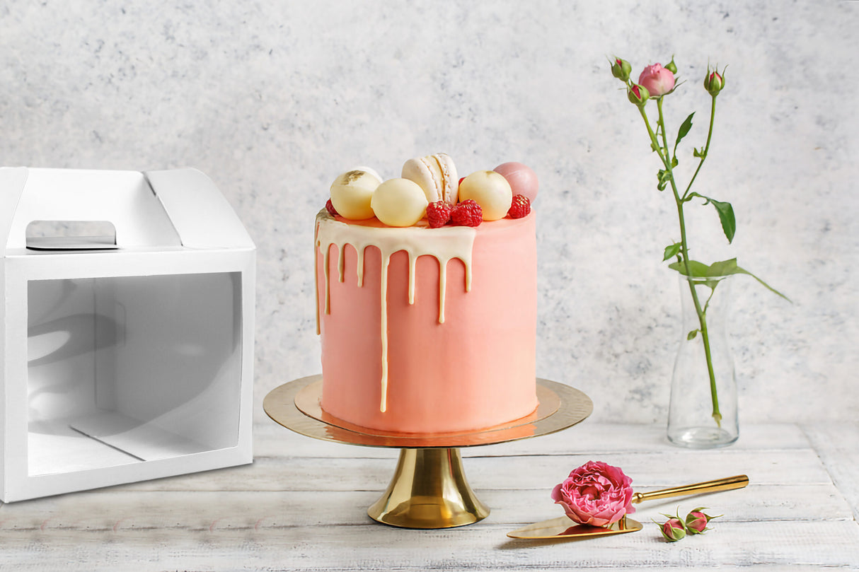 Pink cake with 10x10x8 white paper cake box with window and handle in the background